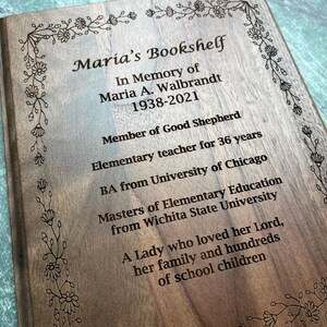 May include: Wooden plaque engraved with the text "Maria's Bookshelf" and an inscription in memory of Maria A. Walbrandt, including her dates of birth and death, and other details. The plaque is framed with a floral border.
