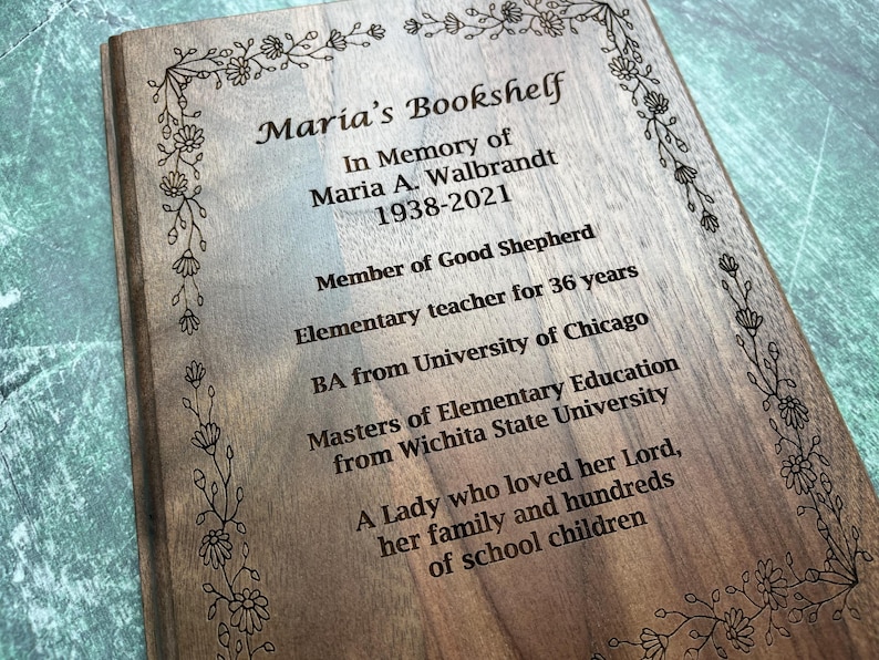 May include: Wooden plaque engraved with the text "Maria's Bookshelf" and an inscription in memory of Maria A. Walbrandt, including her dates of birth and death, and other details. The plaque is framed with a floral border.