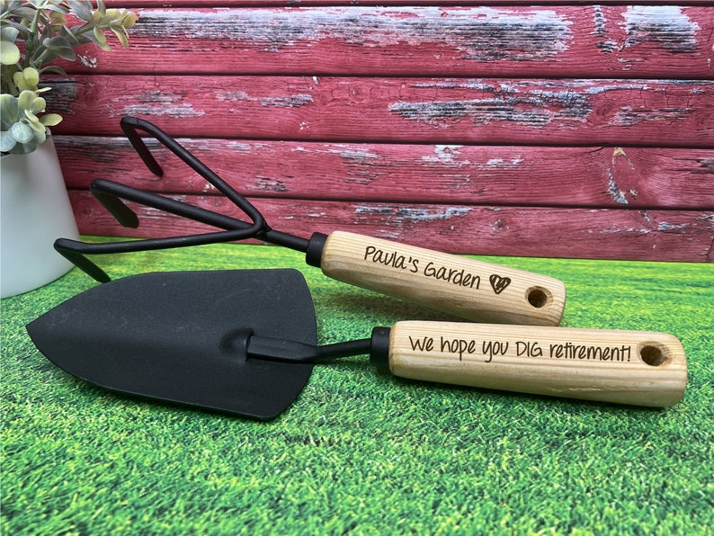 May include: A set of two gardening tools with wooden handles. The handle of the trowel is engraved with the text "We hope you DIG retirement!" and the handle of the cultivator is engraved with the text "Paula's Garden" and a heart symbol.