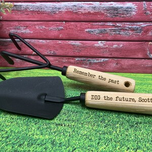 May include: A set of two gardening tools with wooden handles. The handle of the trowel is engraved with the text "DIG the future, Scott!" and the handle of the cultivator is engraved with the text "Remember the past".