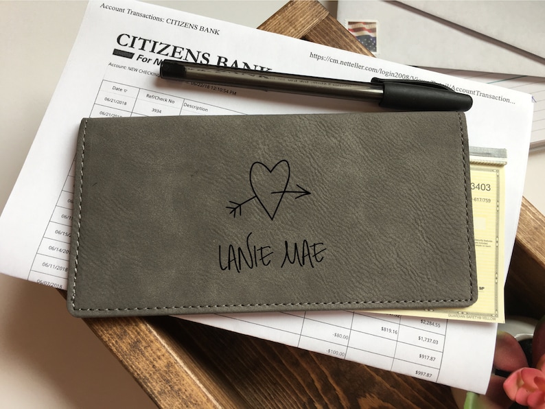 May include: A rectangular gray checkbook cover with a textured finish, featuring a black line drawing of a heart pierced by an arrow and the name LANIE MAE printed in black text. The cover is stitched along the edges with matching gray thread.