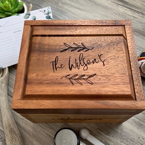 May include: A square wooden recipe box with a hinged lid featuring the engraved text "The Wilson's" flanked by two leaf sprigs. The box has a natural wood grain finish and is positioned alongside a recipe card, a wooden spoon, and small ceramic figurines.