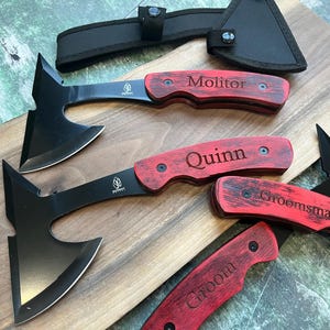 May include: Four personalized black handled axes with red wooden handles. The handles are engraved with the names "Groom", "Groomsman", "Quinn", and "Molitor".