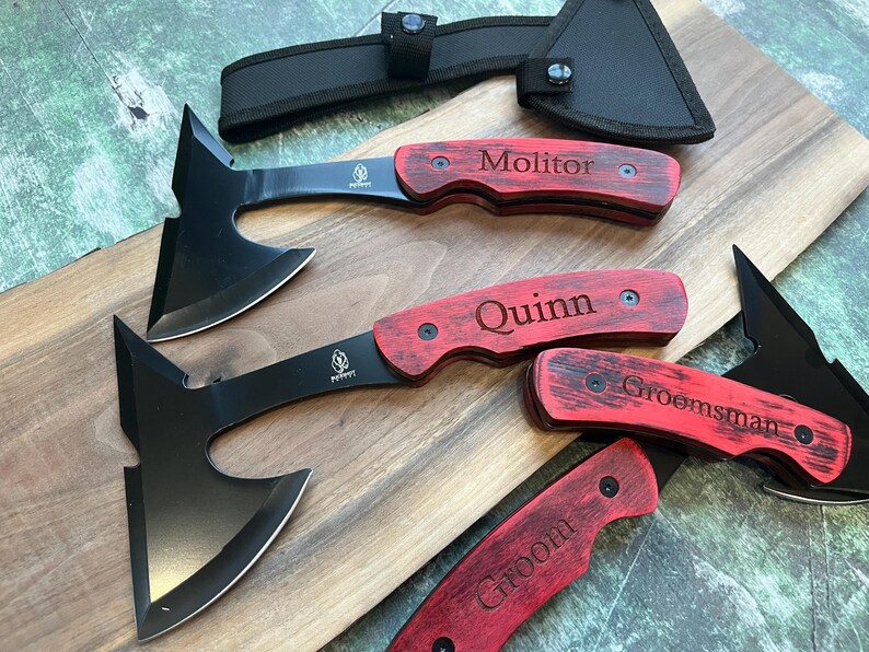 May include: Four personalized black handled axes with red wooden handles. The handles are engraved with the names "Groom", "Groomsman", "Quinn", and "Molitor".