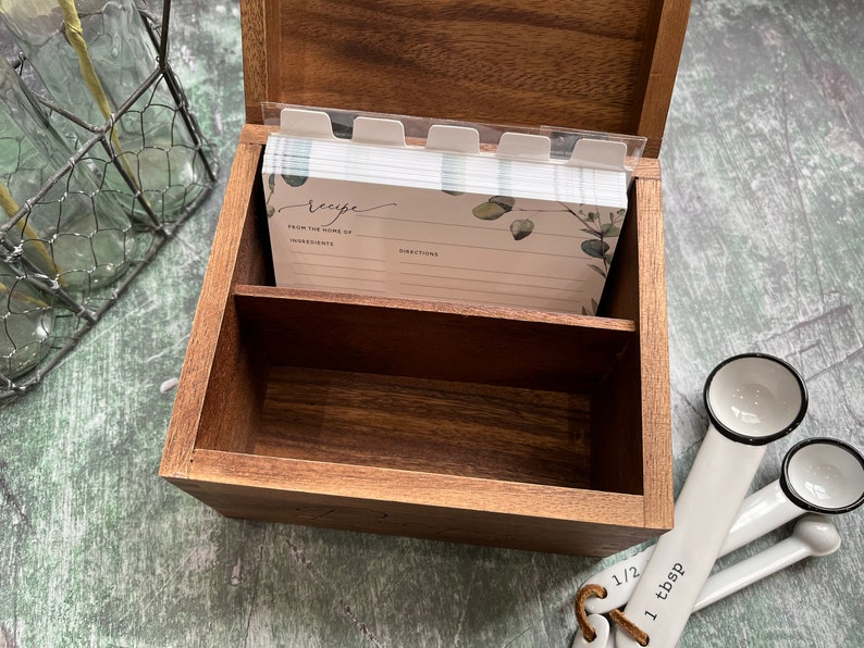 May include: A dark brown wooden recipe box with an open lid containing a stack of white recipe cards featuring eucalyptus leaf illustrations and the text "recipe," "from the home of," "ingredients," and "directions," alongside white measuring spoons.