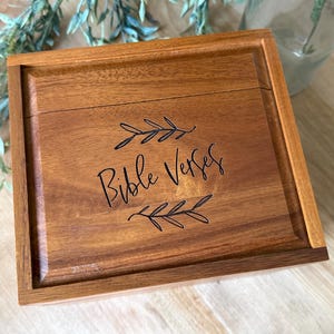 May include: A white ceramic mug with a black handle and the text "But First, Coffee" in a bold, black font. The mug is set against a blurred background, suggesting a cozy setting.