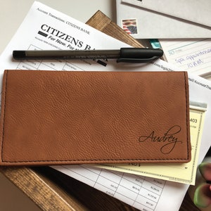 May include: A rectangular tan checkbook cover with a textured surface and black stitching along the edges. The name Audrey is engraved in a black cursive script on the bottom right corner of the cover. A black pen rests on top of the cover.