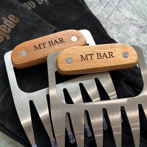May include: Two stainless steel meat shredder claws with wooden handles. The handles are engraved with "MT BAR" and secured with silver rivets. The claws are resting on a black surface.