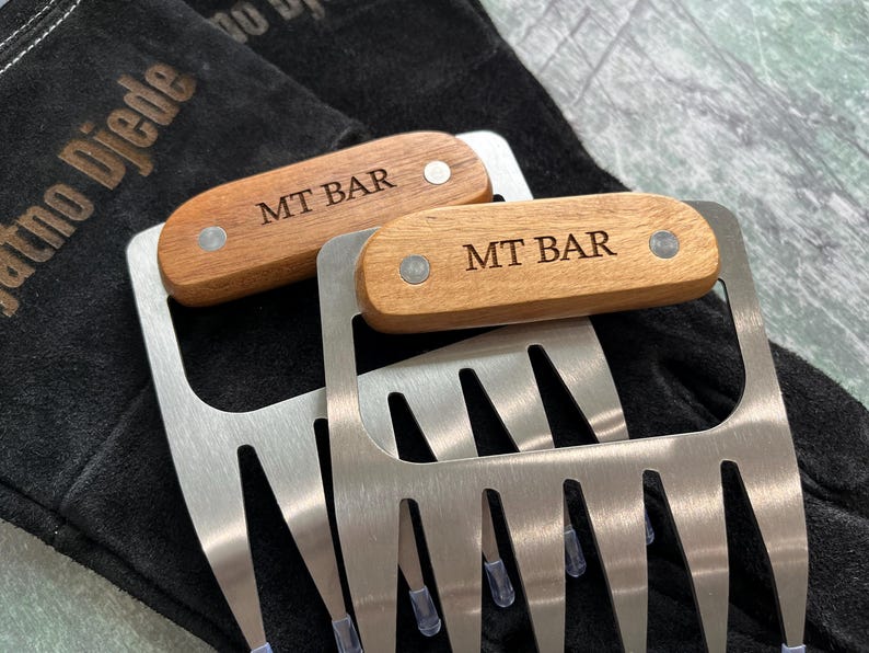 May include: Two stainless steel meat shredder claws with wooden handles. The handles are engraved with "MT BAR" and secured with silver rivets. The claws are resting on a black surface.