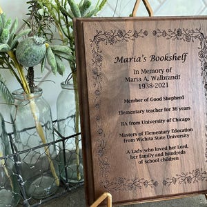 May include: A wooden plaque with the inscription "Maria's Bookshelf" and an obituary for Maria A. Walbrandt, 1938-2021. The plaque is framed with floral designs and is displayed on a gold stand. The background includes glass bottles and greenery.
