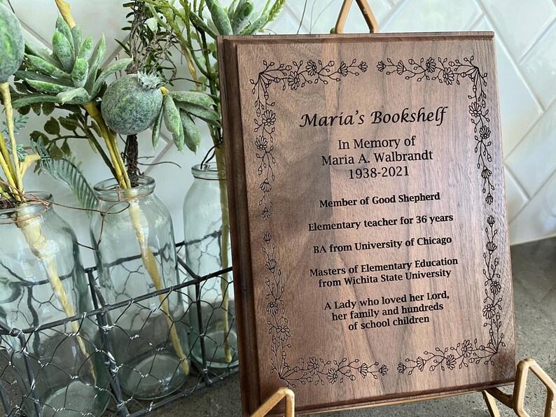 May include: A wooden plaque with the inscription "Maria's Bookshelf" and an obituary for Maria A. Walbrandt, 1938-2021. The plaque is framed with floral designs and is displayed on a gold stand. The background includes glass bottles and greenery.