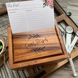 May include: A wooden recipe box with a hinged lid engraved with the text Sara's Recipes and leaf motifs. A white recipe card with floral accents is tucked into the lid slot. Three white measuring spoons are attached to the side of the box.