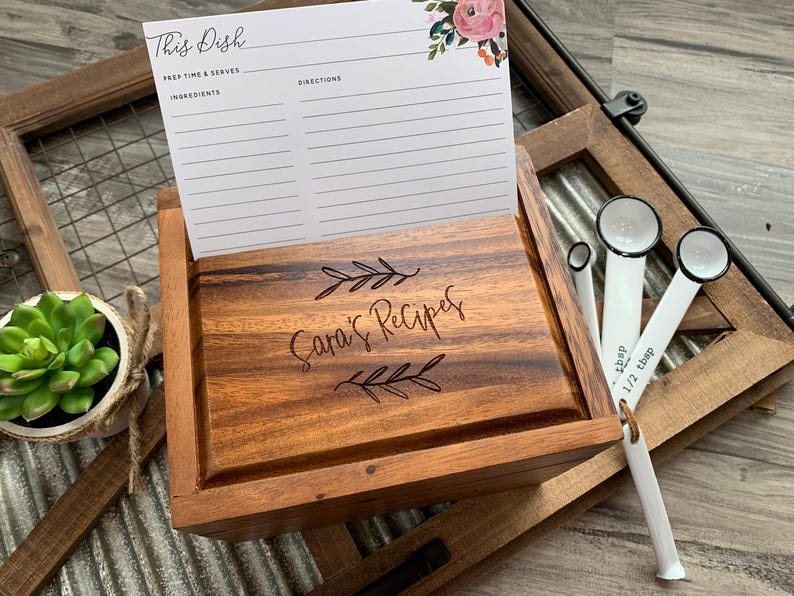 May include: A wooden recipe box with a hinged lid engraved with the text Sara's Recipes and leaf motifs. A white recipe card with floral accents is tucked into the lid slot. Three white measuring spoons are attached to the side of the box.