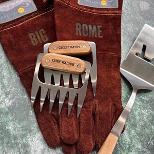 K&ouml;nnte beinhalten: Ein Grillset mit braunen Lederhandschuhen mit den Aufdrucken "BIG" und "ROME", zwei Fleischkrallen aus Edelstahl mit Holzgriffen mit den Gravuren "CHEF DADDY" und "CHEF WALTER" sowie einem Edelstahl-Pfannenwender.