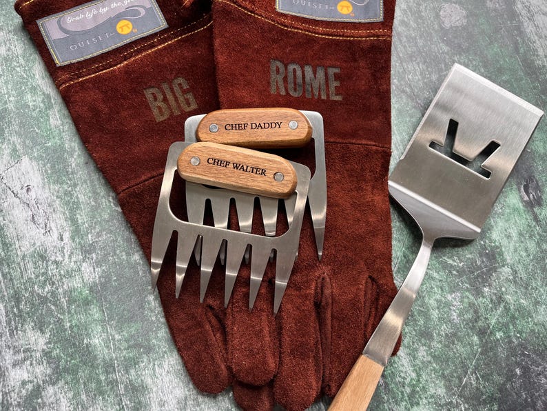 K&ouml;nnte beinhalten: Ein Grillset mit braunen Lederhandschuhen mit den Aufdrucken "BIG" und "ROME", zwei Fleischkrallen aus Edelstahl mit Holzgriffen mit den Gravuren "CHEF DADDY" und "CHEF WALTER" sowie einem Edelstahl-Pfannenwender.
