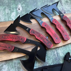 May include: Five black and silver personalized hatchets with red wooden handles. The handles are engraved with the names "Groomsman", "Usher", "Ryan", "Quinn", and "Mollitor".