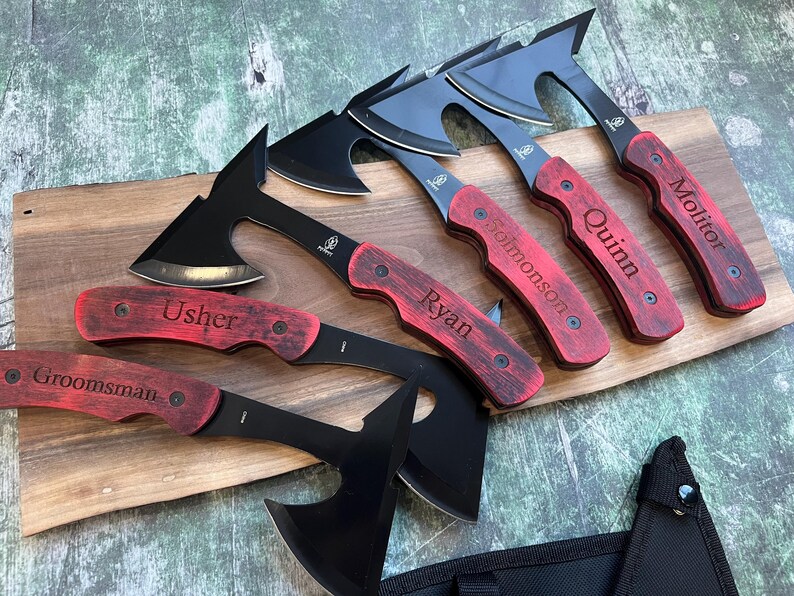 May include: Five black and silver personalized hatchets with red wooden handles. The handles are engraved with the names "Groomsman", "Usher", "Ryan", "Quinn", and "Mollitor".
