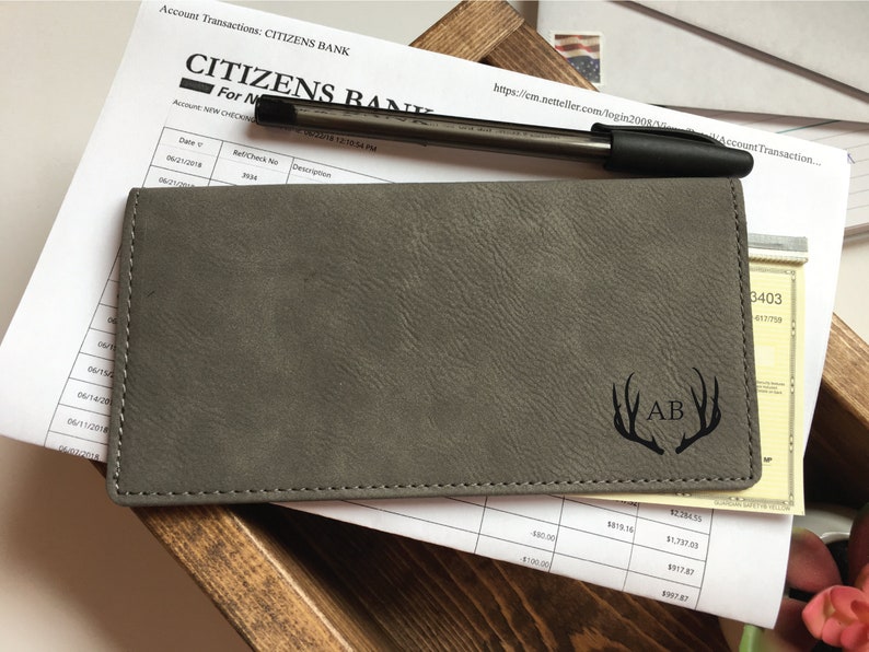 Puede incluir: Una funda rectangular gris para chequera con superficie texturizada, que presenta un dise&ntilde;o grabado con l&aacute;ser de dos astas de ciervo flanqueando las iniciales AB. La funda est&aacute; cosida a lo largo de los bordes y descansa sobre un documento bancario.