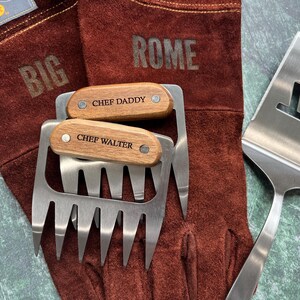 K&ouml;nnte beinhalten: Ein Set Grillwerkzeuge, darunter zwei Fleischkrallen mit Holzgriffen mit den Gravuren "CHEF DADDY" und "CHEF WALTER", ein Pfannenwender und ein Paar braune Leder-Grillhandschuhe. Die Handschuhe sind mit den Worten "BIG" und "ROME" gepr&auml;gt.
