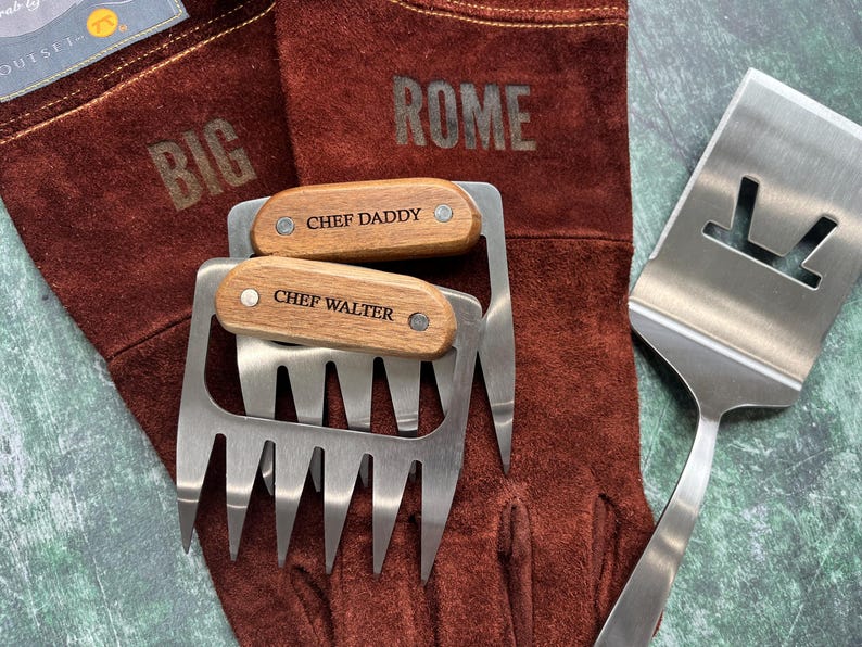 K&ouml;nnte beinhalten: Ein Set Grillwerkzeuge, darunter zwei Fleischkrallen mit Holzgriffen mit den Gravuren "CHEF DADDY" und "CHEF WALTER", ein Pfannenwender und ein Paar braune Leder-Grillhandschuhe. Die Handschuhe sind mit den Worten "BIG" und "ROME" gepr&auml;gt.