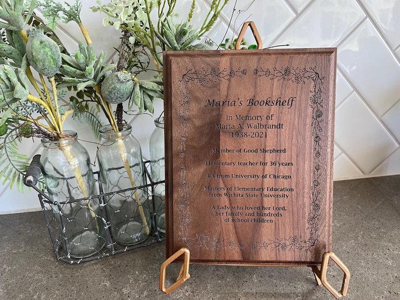 May include: A wooden memorial plaque with the text "Maria's Bookshelf" and an inscription honoring Maria A. Walbrandt, including her birth and death years, and other details. The plaque is displayed on a gold-colored stand.
