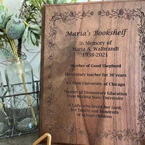 May include: A wooden plaque with the inscription "Maria's Bookshelf" and a floral border. The plaque commemorates Maria A. Walbrandt, including her birth and death years, and her career as an elementary teacher for 36 years.