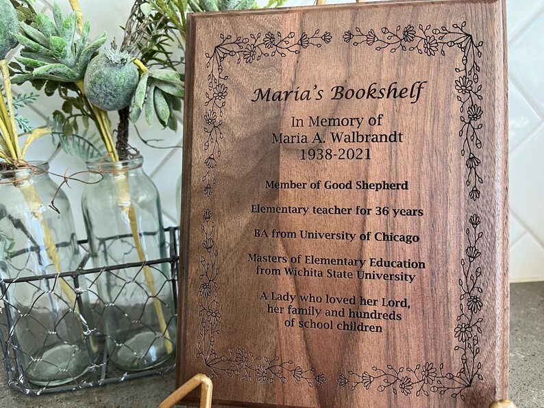 May include: A wooden plaque with the inscription "Maria's Bookshelf" and a floral border. The plaque commemorates Maria A. Walbrandt, including her birth and death years, and her career as an elementary teacher for 36 years.