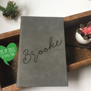 Puede incluir: Un diario de imitaci&oacute;n de cuero gris con el nombre "Brooke" escrito en cursiva negra. El diario est&aacute; en una caja de madera con una peque&ntilde;a planta suculenta en maceta y un bol&iacute;grafo negro.