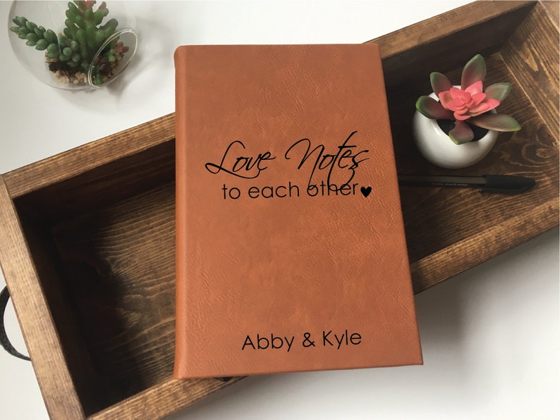 Op de afbeelding: Een bruin lederen gebonden dagboek met de tekst "Love Notes to each other" en "Abby & Kyle" gegraveerd op de voorzijde. Het dagboek ligt in een houten doos.