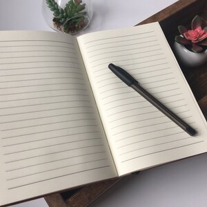 Puede incluir: Un cuaderno de tapa dura abierto con un bol&iacute;grafo negro sobre la p&aacute;gina derecha. El cuaderno est&aacute; apoyado en una caja de madera con una peque&ntilde;a planta suculenta en maceta.