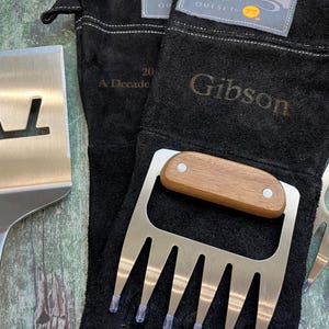 K&ouml;nnte beinhalten: Ein Edelstahl-Pfannenwender mit einem Ausschnitt und ein Satz Edelstahl-Fleischkrallen mit Holzgriffen werden mit schwarzen Grillhandschuhen pr&auml;sentiert. Auf den Handschuhen steht das Wort "Gibson".