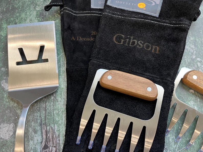K&ouml;nnte beinhalten: Ein Edelstahl-Pfannenwender mit einem Ausschnitt und ein Satz Edelstahl-Fleischkrallen mit Holzgriffen werden mit schwarzen Grillhandschuhen pr&auml;sentiert. Auf den Handschuhen steht das Wort "Gibson".