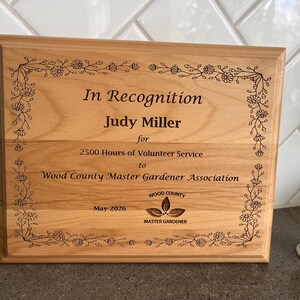 May include: A rectangular wooden plaque with the text "In Recognition Judy Miller" and other text. The plaque is framed with a floral design. The plaque is for 2500 hours of volunteer service to the Wood County Master Gardener Association. The date is May 2026.
