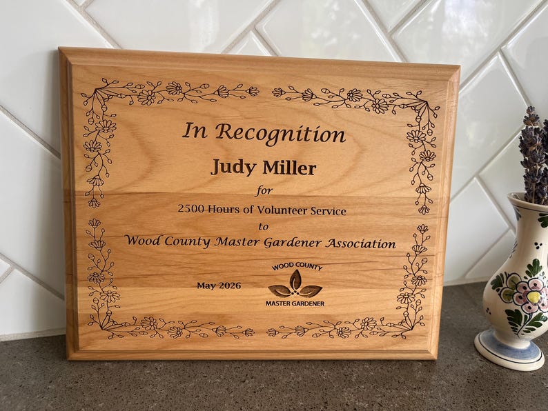 May include: A rectangular wooden plaque with the text "In Recognition Judy Miller" and other text. The plaque is framed with a floral design. The plaque is for 2500 hours of volunteer service to the Wood County Master Gardener Association. The date is May 2026.