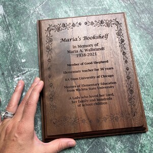 May include: A wooden plaque with engraved text and floral border. The text reads "Maria's Bookshelf" and includes an inscription in memory of Maria A. Walbrandt, 1938-2021, detailing her life as a teacher and her love for her family.