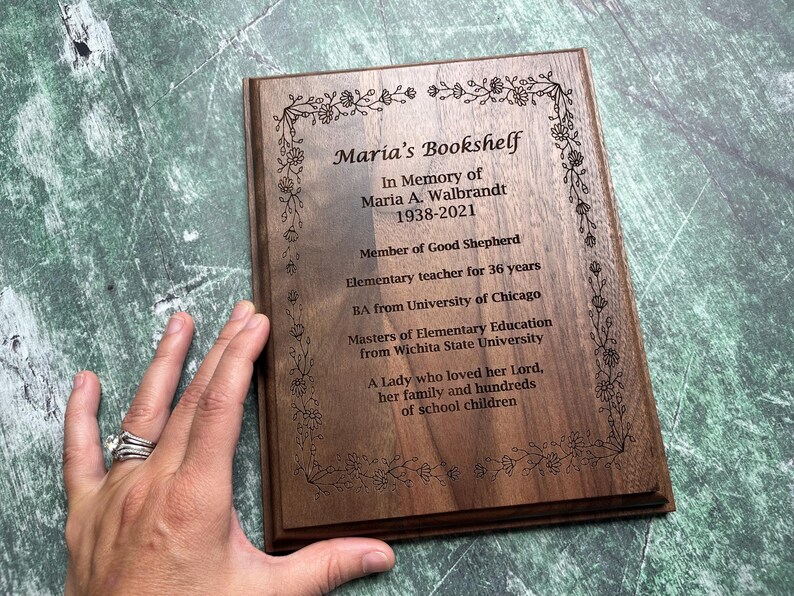 May include: A wooden plaque with engraved text and floral border. The text reads "Maria's Bookshelf" and includes an inscription in memory of Maria A. Walbrandt, 1938-2021, detailing her life as a teacher and her love for her family.