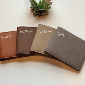 May include: Four rectangular bi-fold wallets arranged in a fan shape. The colors from left to right are labeled as Rawhide, Dk. Brown, Lt. Brown, and Gray. Each wallet features a smooth, solid-colored exterior with visible stitching along the edges.