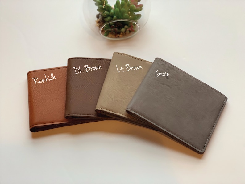 May include: Four rectangular bi-fold wallets arranged in a fan shape. The colors from left to right are labeled as Rawhide, Dk. Brown, Lt. Brown, and Gray. Each wallet features a smooth, solid-colored exterior with visible stitching along the edges.