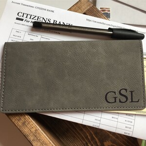 May include: A rectangular grey checkbook cover with a textured surface and the letters GSL embossed in black on the bottom right corner. The cover is positioned over a Citizens Bank account transaction statement and a black ballpoint pen.