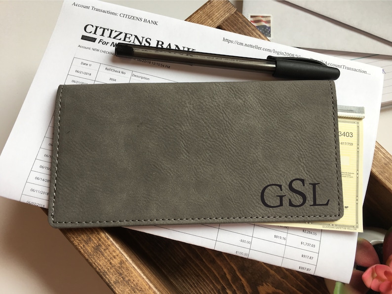 May include: A rectangular grey checkbook cover with a textured surface and the letters GSL embossed in black on the bottom right corner. The cover is positioned over a Citizens Bank account transaction statement and a black ballpoint pen.