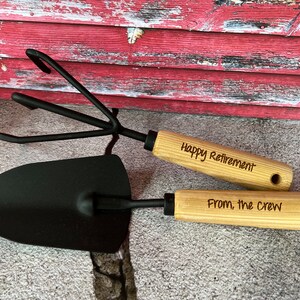 May include: A set of two gardening tools with wooden handles. The handles are engraved with the text "Happy Retirement" and "From, the crew".