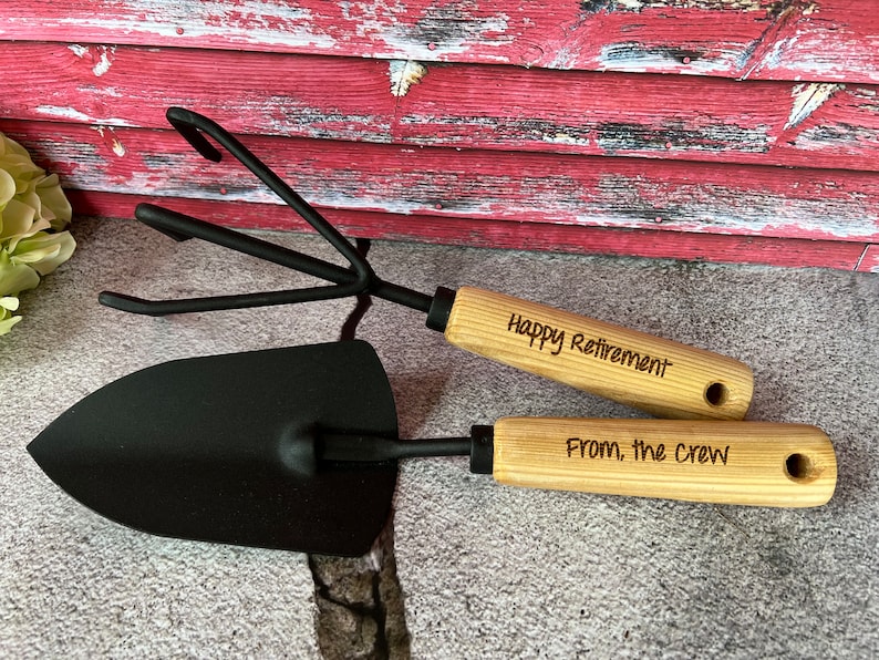 May include: A set of two gardening tools with wooden handles. The handles are engraved with the text "Happy Retirement" and "From, the crew".