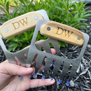 May include: Two stainless steel meat shredder claws with wooden handles. The handles are engraved with the initials "DW". The claws have four tines each and are designed for shredding cooked meats. The handles are attached with silver rivets.