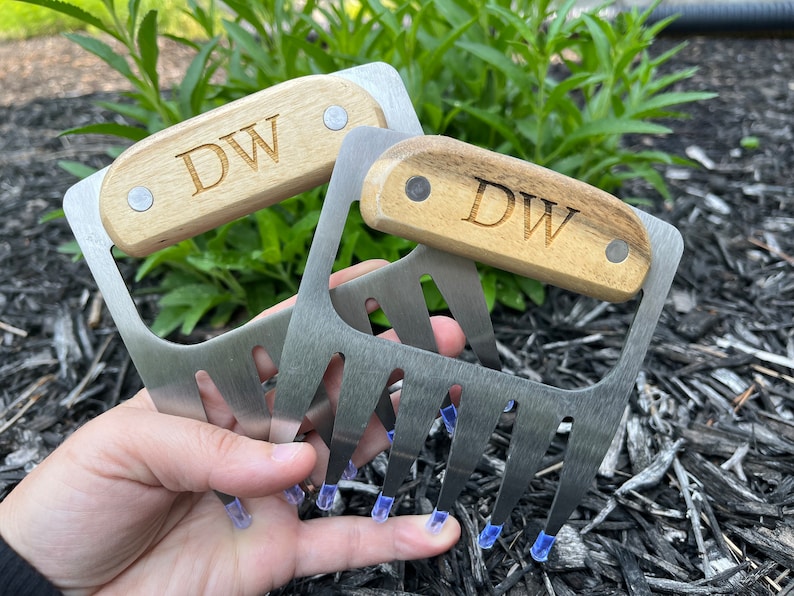 May include: Two stainless steel meat shredder claws with wooden handles. The handles are engraved with the initials "DW". The claws have four tines each and are designed for shredding cooked meats. The handles are attached with silver rivets.