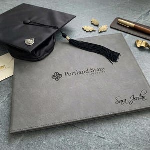 May include: A gray diploma cover featuring the Portland State University logo and the name Sara Jordan, accompanied by a black graduation cap with a tassel, a cream envelope, three gold leaf pins, a brown pen, and a gold metal object.