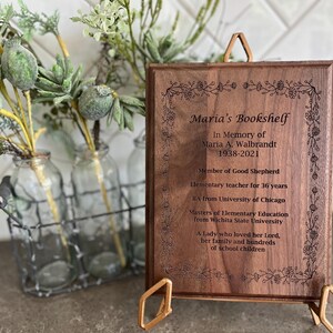 May include: A wooden plaque with the inscription "Maria's Bookshelf" and an obituary. The plaque is supported by a gold-colored stand. Decorative floral elements are etched around the border. A wire basket with glass bottles and greenery is in the background.