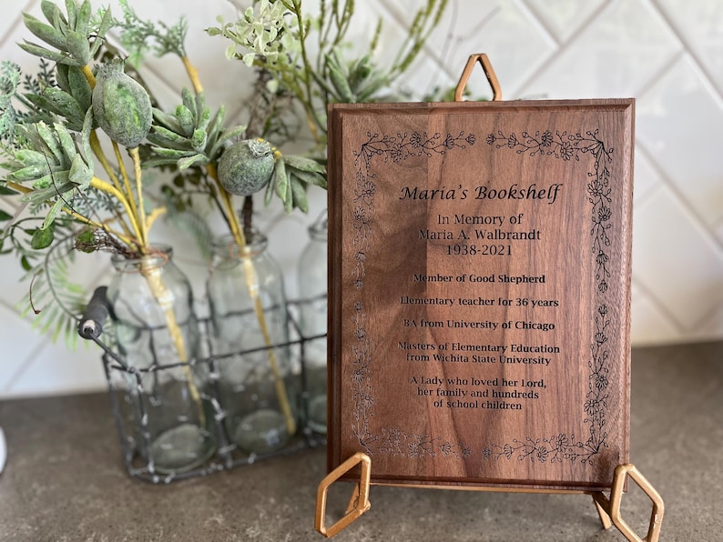 May include: A wooden plaque with the inscription "Maria's Bookshelf" and an obituary. The plaque is supported by a gold-colored stand. Decorative floral elements are etched around the border. A wire basket with glass bottles and greenery is in the background.