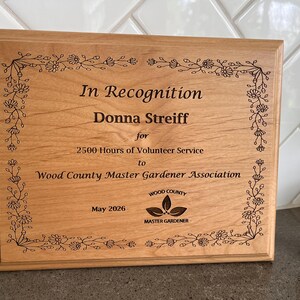 May include: A wooden plaque with the text "In Recognition" and the name "Donna Streiff". The plaque is for 2500 hours of volunteer service to the Wood County Master Gardener Association. The plaque is framed with a floral design.