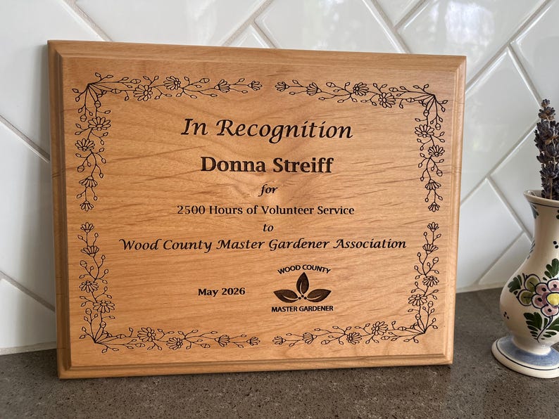 May include: A wooden plaque with the text "In Recognition" and the name "Donna Streiff". The plaque is for 2500 hours of volunteer service to the Wood County Master Gardener Association. The plaque is framed with a floral design.