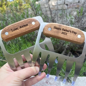 May include: Two stainless steel meat shredder claws with wooden handles. One claw is engraved with "Love, Archer", the other with "Happy Father's Day 2023". The claws have blue plastic tips.